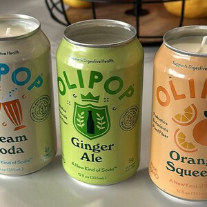 May include: Three 12-ounce aluminum cans of Olipop sparkling probiotic soda. The flavors are Cream Soda, Ginger Ale, and Orange Squeeze. Each can has a different color and design.