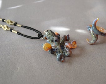 May include: A glass octopus pendant necklace and ring set. The octopus is a dark blue and green color with orange accents. The necklace has a black cord and gold chain.