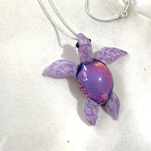 Könnte beinhalten: Ein violetter gläserner Meeresschildkröten-Anhänger an einer silbernen Kette. Die Schildkröte hat eine irisierende Oberfläche und detaillierte Flügel.