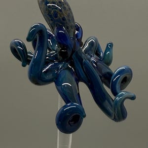 May include: A handcrafted glass octopus sculpture in shades of blue. The octopus has eight curled tentacles and a detailed head with a textured pattern. The sculpture is mounted on a clear stand with a teal base.
