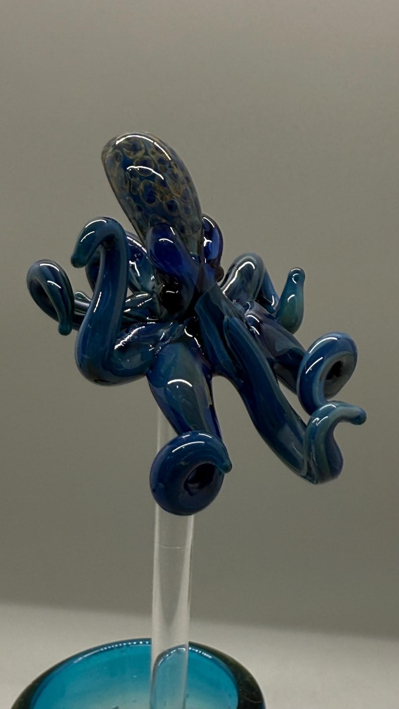 May include: A handcrafted glass octopus sculpture in shades of blue. The octopus has eight curled tentacles and a detailed head with a textured pattern. The sculpture is mounted on a clear stand with a teal base.