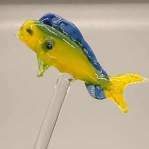 Puede incluir: Figura de pez de cristal colorido con tonos amarillos, azules y verdes. El pez está montado sobre una varilla de cristal transparente, mostrando su diseño vibrante. La cola y las aletas de la figura son detalladas.