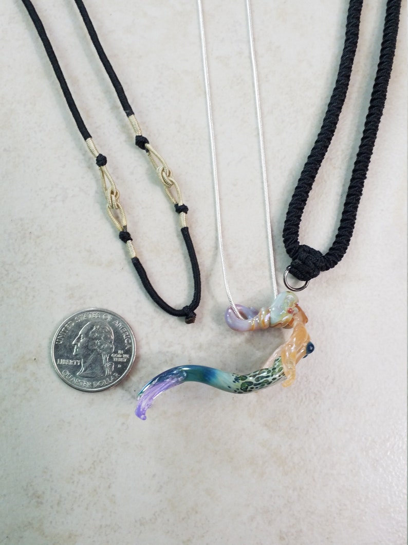Pode incluir: Tr&ecirc;s colares com pingentes exclusivos. Um tem um cord&atilde;o preto com detalhes dourados, outro tem uma corrente prateada e o terceiro tem um cord&atilde;o preto. Os pingentes s&atilde;o coloridos, com um design abstrato curvo. Uma moeda est&aacute; inclu&iacute;da para refer&ecirc;ncia de tamanho.