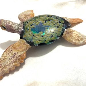 May include: A glass sea turtle sculpture with a green, blue, and black shell and a light brown body. The turtle has a detailed, textured surface.