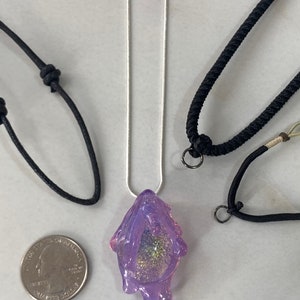 May include: A purple resin pendant with glitter and a dripping effect, hanging from a silver chain. The pendant is displayed on a white surface with two black cords.