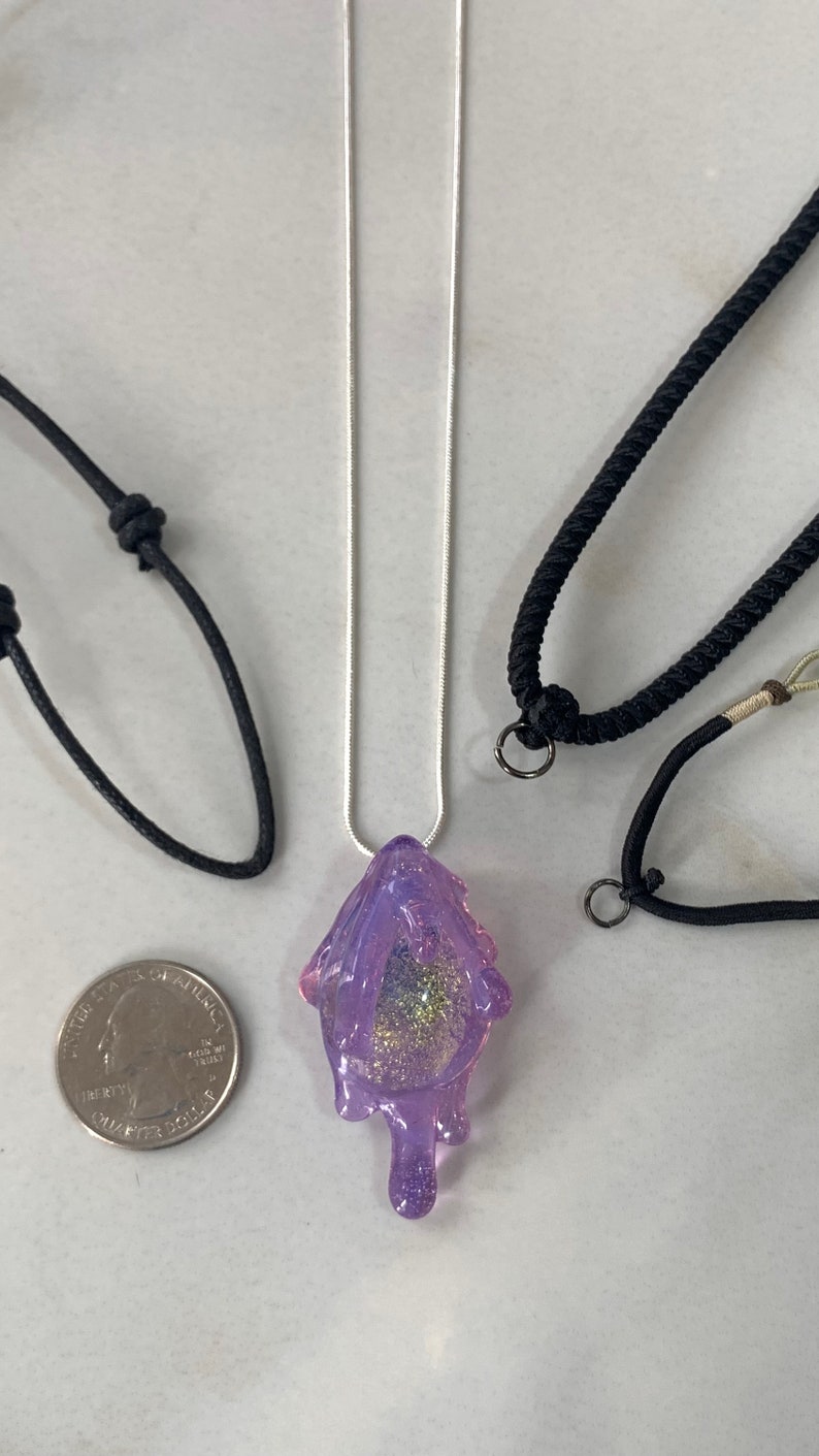 May include: A purple resin pendant with glitter and a dripping effect, hanging from a silver chain. The pendant is displayed on a white surface with two black cords.