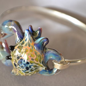 May include: A silver bangle bracelet with a colorful glass crab charm. The crab is blue, green, and yellow with black eyes.