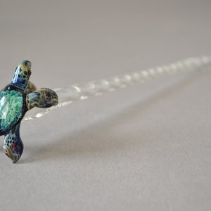 Pode incluir: Uma figurinha de tartaruga marinha de vidro com detalhes pintados em azul, verde e dourado. A tartaruga está pousada em uma haste de vidro transparente.