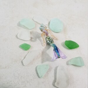 Pode incluir: Uma estatueta de sereia de vidro com cauda roxa e azul, torso com padr&atilde;o verde e branco e cabelo loiro. A sereia &eacute; cercada por vidro do mar em tons de verde e branco. As pe&ccedil;as de vidro do mar s&atilde;o de v&aacute;rias formas e tamanhos.
