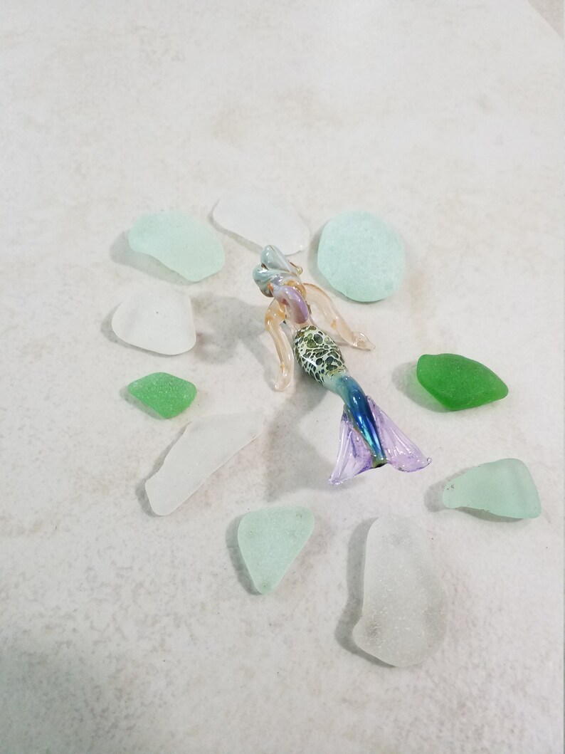 Pode incluir: Uma estatueta de sereia de vidro com cauda roxa e azul, torso com padr&atilde;o verde e branco e cabelo loiro. A sereia &eacute; cercada por vidro do mar em tons de verde e branco. As pe&ccedil;as de vidro do mar s&atilde;o de v&aacute;rias formas e tamanhos.