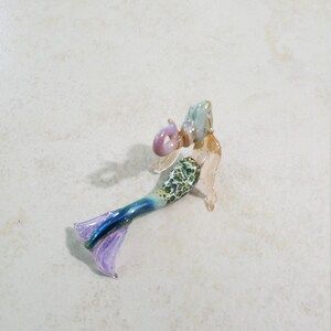 Pode incluir: Pequena estatueta de sereia de vidro. A sereia tem uma cabe&ccedil;a azul clara e rosa, um torso com padr&atilde;o verde e branco e uma cauda azul e roxa. A estatueta est&aacute; sobre uma superf&iacute;cie clara.