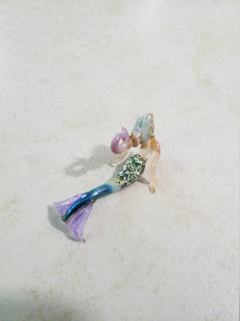 Pode incluir: Pequena estatueta de sereia de vidro. A sereia tem uma cabe&ccedil;a azul clara e rosa, um torso com padr&atilde;o verde e branco e uma cauda azul e roxa. A estatueta est&aacute; sobre uma superf&iacute;cie clara.