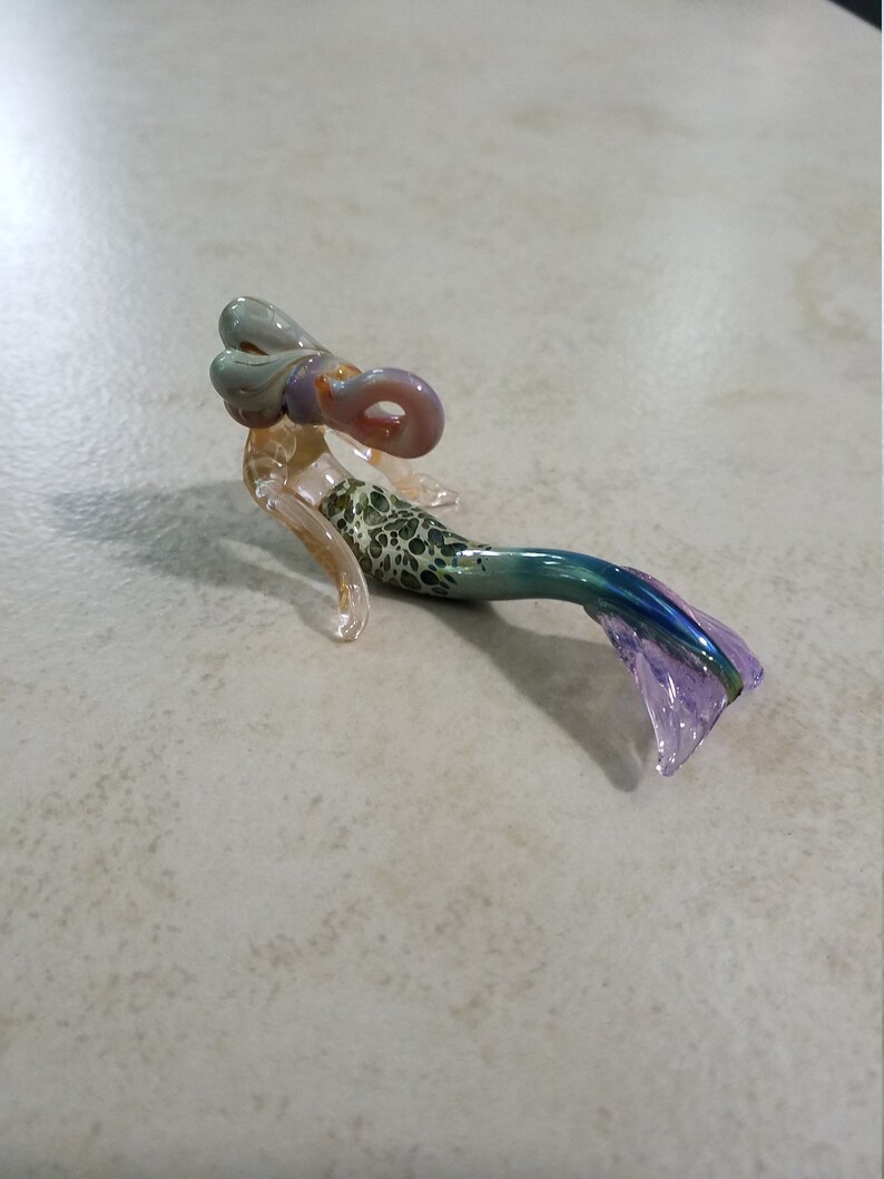 Pode incluir: Uma estatueta de sereia de vidro artesanal. A sereia tem uma cauda manchada de verde e branco, uma barbatana azul e roxa e um corpo de cor clara. O cabelo da sereia &eacute; uma mistura de verde claro e roxo.
