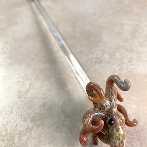May include: A clear glass octopus-shaped dabber tool with a long, thin handle. The octopus is brown, tan, and white with a black eye. The handle has a small, round, amber-colored tip. This unique glass art piece is ideal for concentrates.