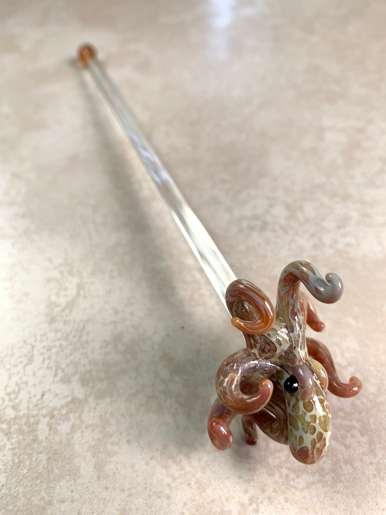May include: A clear glass octopus-shaped dabber tool with a long, thin handle. The octopus is brown, tan, and white with a black eye. The handle has a small, round, amber-colored tip. This unique glass art piece is ideal for concentrates.