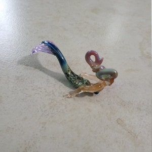 Pode incluir: Pequena estatueta de sereia de vidro. A sereia tem uma cauda azul e roxa, um corpo com padr&atilde;o verde e castanho e um torso e cabe&ccedil;a castanho claro. A sereia est&aacute; sobre uma superf&iacute;cie clara.
