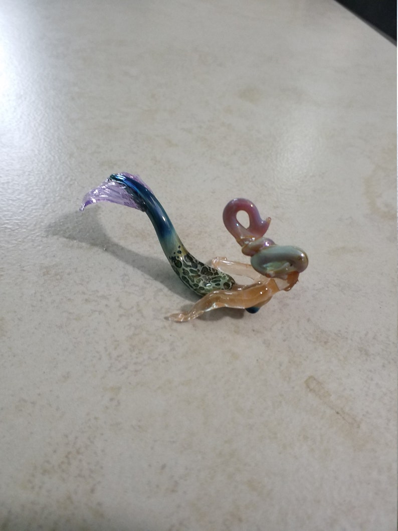 Pode incluir: Pequena estatueta de sereia de vidro. A sereia tem uma cauda azul e roxa, um corpo com padr&atilde;o verde e castanho e um torso e cabe&ccedil;a castanho claro. A sereia est&aacute; sobre uma superf&iacute;cie clara.