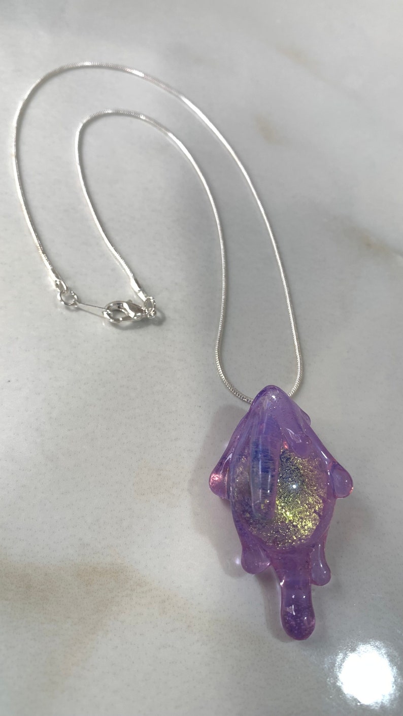 May include: A silver chain necklace with a purple glass pendant. The pendant is shaped like a blob with gold glitter inside.