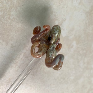 May include: A handcrafted glass octopus sculpture in shades of brown, tan, and purple, with a black eye. The octopus is mounted on a clear glass rod. The sculpture is a unique decorative item.