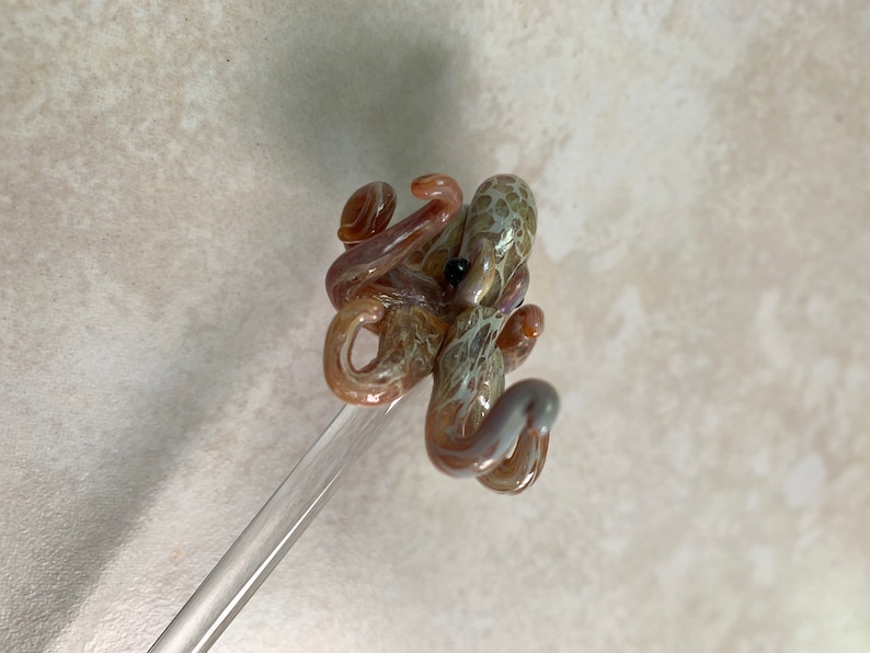 May include: A handcrafted glass octopus sculpture in shades of brown, tan, and purple, with a black eye. The octopus is mounted on a clear glass rod. The sculpture is a unique decorative item.