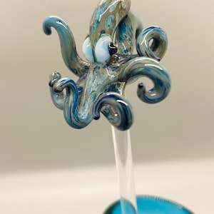 May include: A handcrafted glass octopus sculpture in shades of blue, teal, and brown, mounted on a clear glass rod. The octopus has detailed tentacles and a textured body. The sculpture is set against a soft, neutral background.