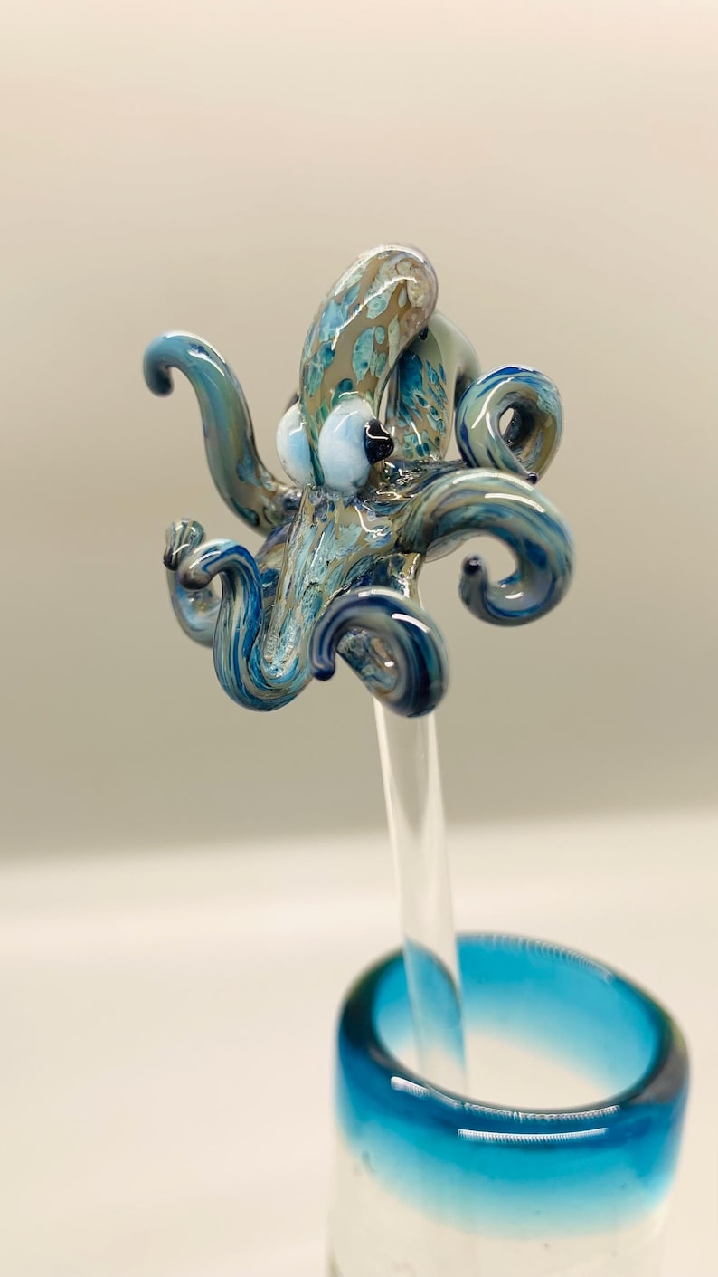 May include: A handcrafted glass octopus sculpture in shades of blue, teal, and brown, mounted on a clear glass rod. The octopus has detailed tentacles and a textured body. The sculpture is set against a soft, neutral background.