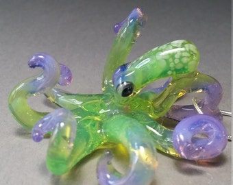 May include: A small glass octopus pendant with green and purple iridescent glass. The octopus has eight tentacles and a single eye.