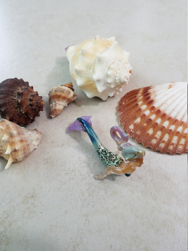 Pode incluir: Uma estatueta de sereia de vidro com cauda azul e roxa, torso verde manchado e pele bronzeada. V&aacute;rias conchas de v&aacute;rios tamanhos e cores circundam a sereia, incluindo uma grande concha de concha creme.