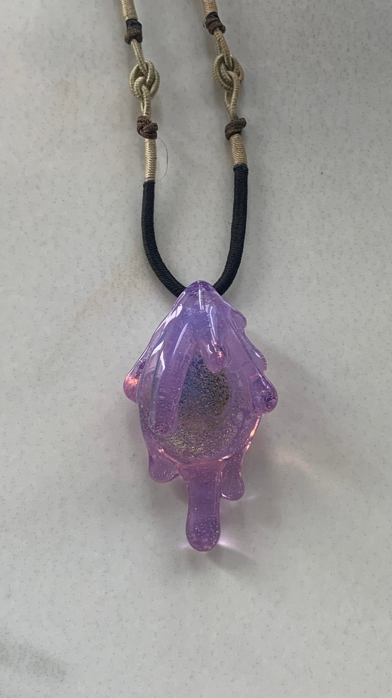 May include: A purple glass pendant necklace with a dripping design. The necklace is made with a black cord and a beige and brown cord.
