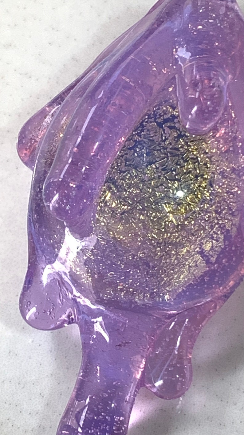May include: A purple glass hand with a sparkly gold and purple interior. The hand is holding a small, round object.
