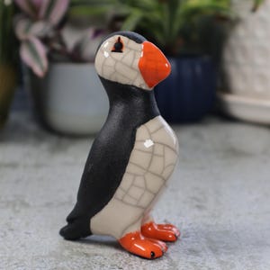 Op de afbeelding: Een keramiek beeldje van een papegaaiduiker met een zwarte kop en een wit lichaam. De papegaaiduiker heeft een oranje snavel en poten. Het beeldje staat op een grijs oppervlak.