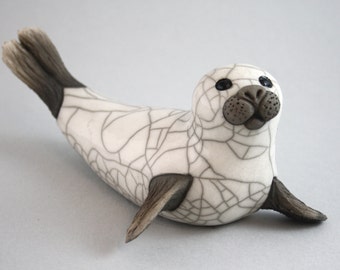 Puede incluir: Una escultura de foca de cerámica blanca con un acabado de esmalte craquelado. La foca tiene ojos negros y una nariz marrón. La escultura está tumbada boca abajo con las aletas extendidas.
