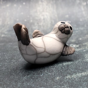 May include: A ceramic seal figurine in a playful pose. The seal is primarily white with dark grey accents on its flippers and tail. The surface has a crackle glaze effect, adding texture. The figurine is approximately 7.6 cm long.