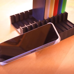 Puede incluir: Un tel&eacute;fono inteligente negro con una pantalla oscura descansa sobre un soporte de pl&aacute;stico negro con m&uacute;ltiples compartimentos. El soporte tiene un dise&ntilde;o a rayas de colores.