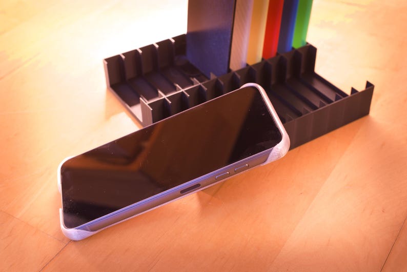 Puede incluir: Un tel&eacute;fono inteligente negro con una pantalla oscura descansa sobre un soporte de pl&aacute;stico negro con m&uacute;ltiples compartimentos. El soporte tiene un dise&ntilde;o a rayas de colores.