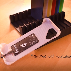 Op de afbeelding: Witte plastic telefoonstandaard met een zwarte oplader en de tekst "Revolution" erop. De standaard staat op een zwart en grijs oppervlak. *Qi-Pad niet inbegrepen*.