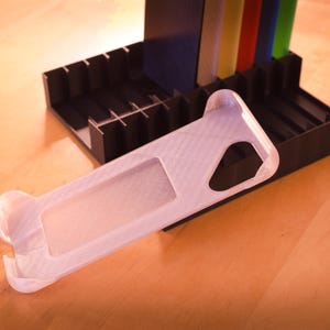 Op de afbeelding: Een witte plastic telefoonhoes met een textuur. De hoes is ontworpen om te passen op een smartphone en heeft een uitsparing voor de camera.