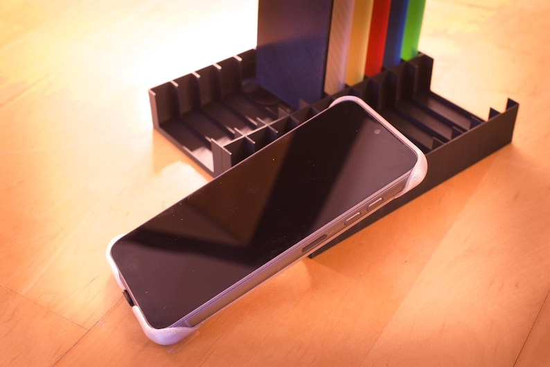 Op de afbeelding: Een zwarte smartphone met een witte hoes ligt op een zwarte plastic standaard. De standaard heeft meerdere sleuven om andere items in op te bergen.