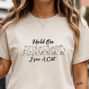 Puede incluir: Camiseta beige con el texto "Hold On I see A Cat" sobre tres caras de gatos de dibujos animados. Los gatos tienen diferentes patrones. La camiseta tiene cuello redondo y mangas cortas.