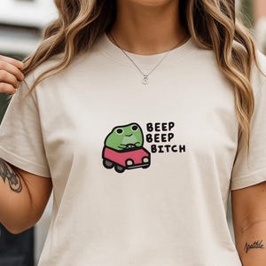 Puede incluir: Camiseta beige con un dibujo de una rana verde sonriente conduciendo un coche rojo. El texto "BEEP BEEP BITCH" está impreso en negro junto a la rana.