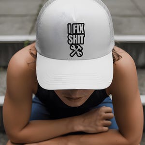 May include: White trucker hat with a mesh back and a curved brim. The front of the hat features the text "I FIX SHIT" in bold black letters, with a hammer and wrench graphic below. The hat is worn by a person.