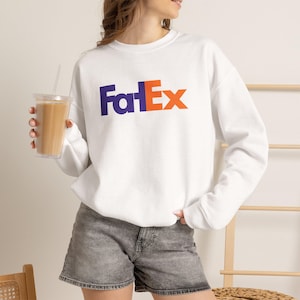 May include: A white long-sleeve crewneck sweatshirt featuring the text "FatEx" printed across the chest in purple and orange lettering. The garment has a standard fit with ribbed cuffs and a ribbed neckline, paired with gray denim shorts.