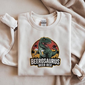Puede incluir: Sudadera color crema con un dinosaurio verde sosteniendo jarras de cerveza. El diseño incluye el texto "BEEROSAURUS BEER-REX" en negro y amarillo, sobre un fondo de puesta de sol con palmeras.