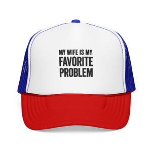Può includere: Un cappellino trucker con pannello frontale bianco con il testo nero MY WIFE IS MY FAVORITE PROBLEM, visiera rossa e pannelli posteriori in rete blu. Il cappello presenta una corona strutturata e un bottone sulla parte superiore.