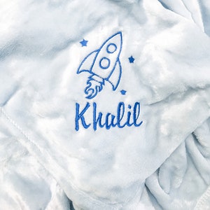 K&ouml;nnte beinhalten: Eine hellblaue Babydecke mit einer wei&szlig;en gestickten Rakete und Sternen. Die Decke ist mit dem Namen "Khalil" personalisiert.