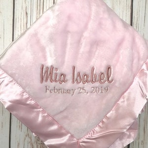 May include: Pink baby blanket with a satin border. The blanket is embroidered with the name "Mia Isabel" and the date "February 25, 2019".