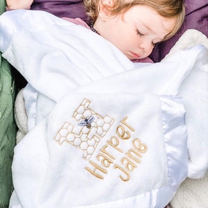 May include: White baby blanket with a satin border. The blanket has a gold embroidered letter H with a bee inside and the name "Harper Jane" embroidered below.