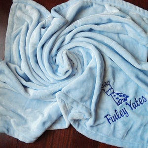 Pode incluir: Um cobertor de beb&ecirc; azul claro com uma pequena girafa bordada e o nome "Finley Yates" em fio azul.
