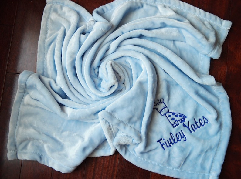 Pode incluir: Um cobertor de beb&ecirc; azul claro com uma pequena girafa bordada e o nome "Finley Yates" em fio azul.