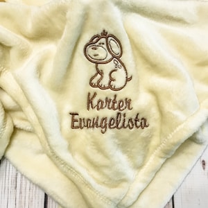 Pode incluir: Um cobertor de pel&uacute;cia de cor creme com um cachorro bordado em marrom e o nome "Karter Evangelista" bordado abaixo.